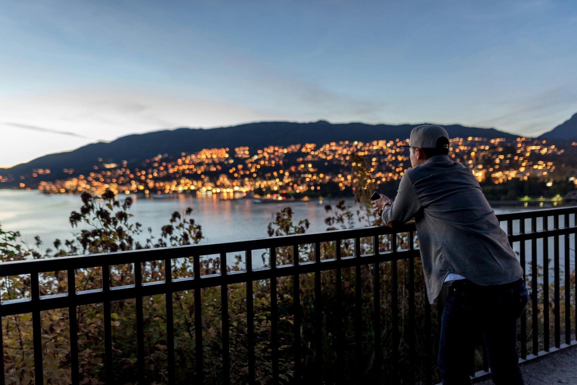 Adult man gazing at illuminated cityscape by the bay at dusk, capturing tranquility and scenic beauty.