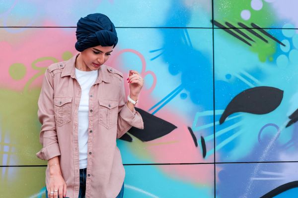 Stylish woman wearing a turban stands before a vibrant graffiti wall, showcasing modern urban fashion.